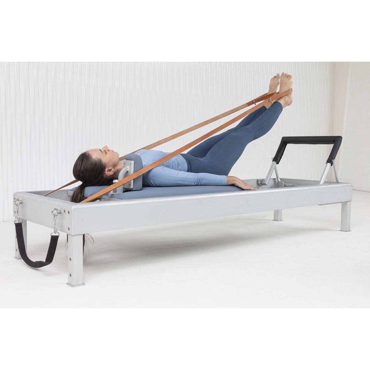 User completing leg strength work with foot straps on the Elina Pilates Classic Aluminum Reformer 86″ in a studio setting.