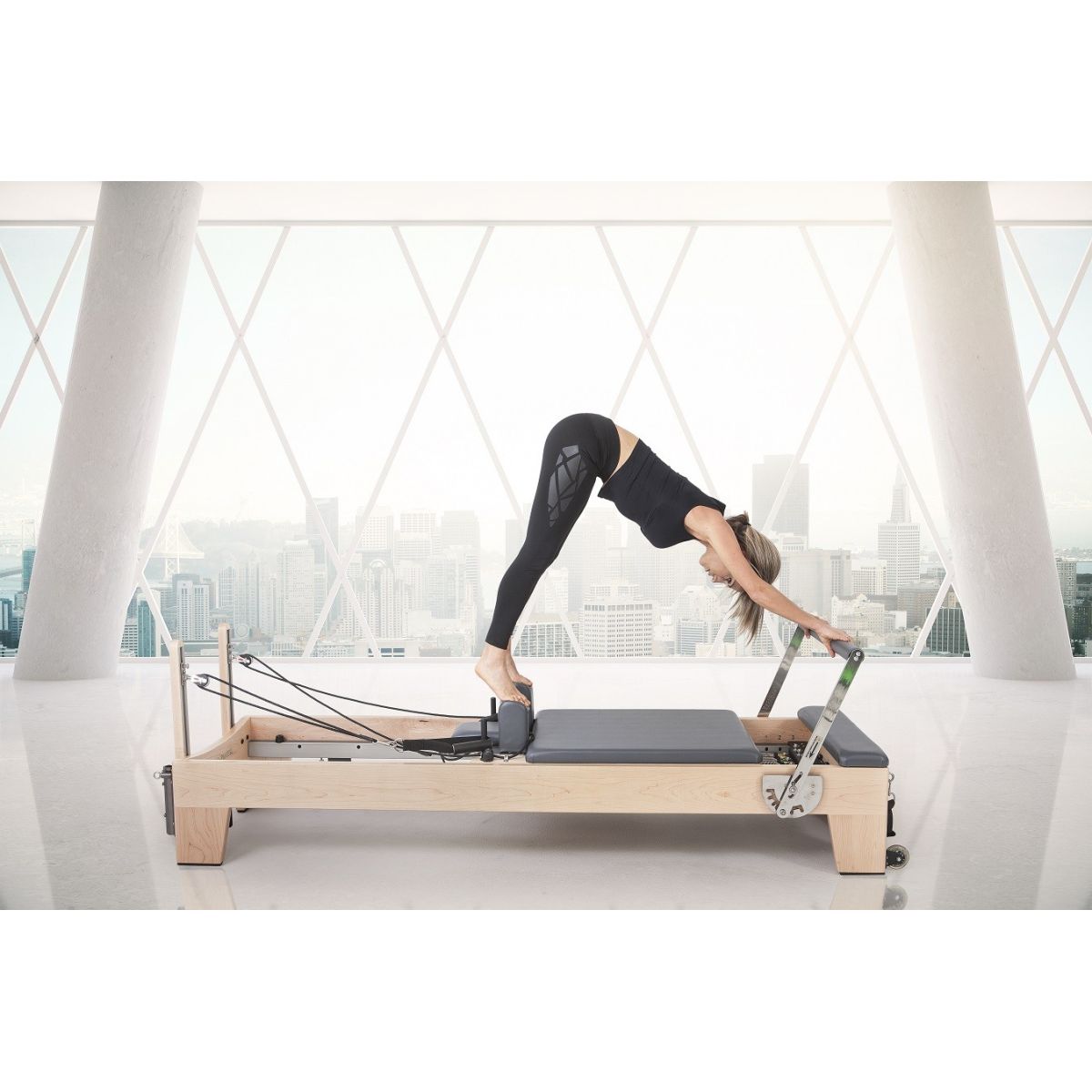 Woman performing tower stretch exercise on Elina Pilates Wood Reformer with Tower Pro in a studio setting.